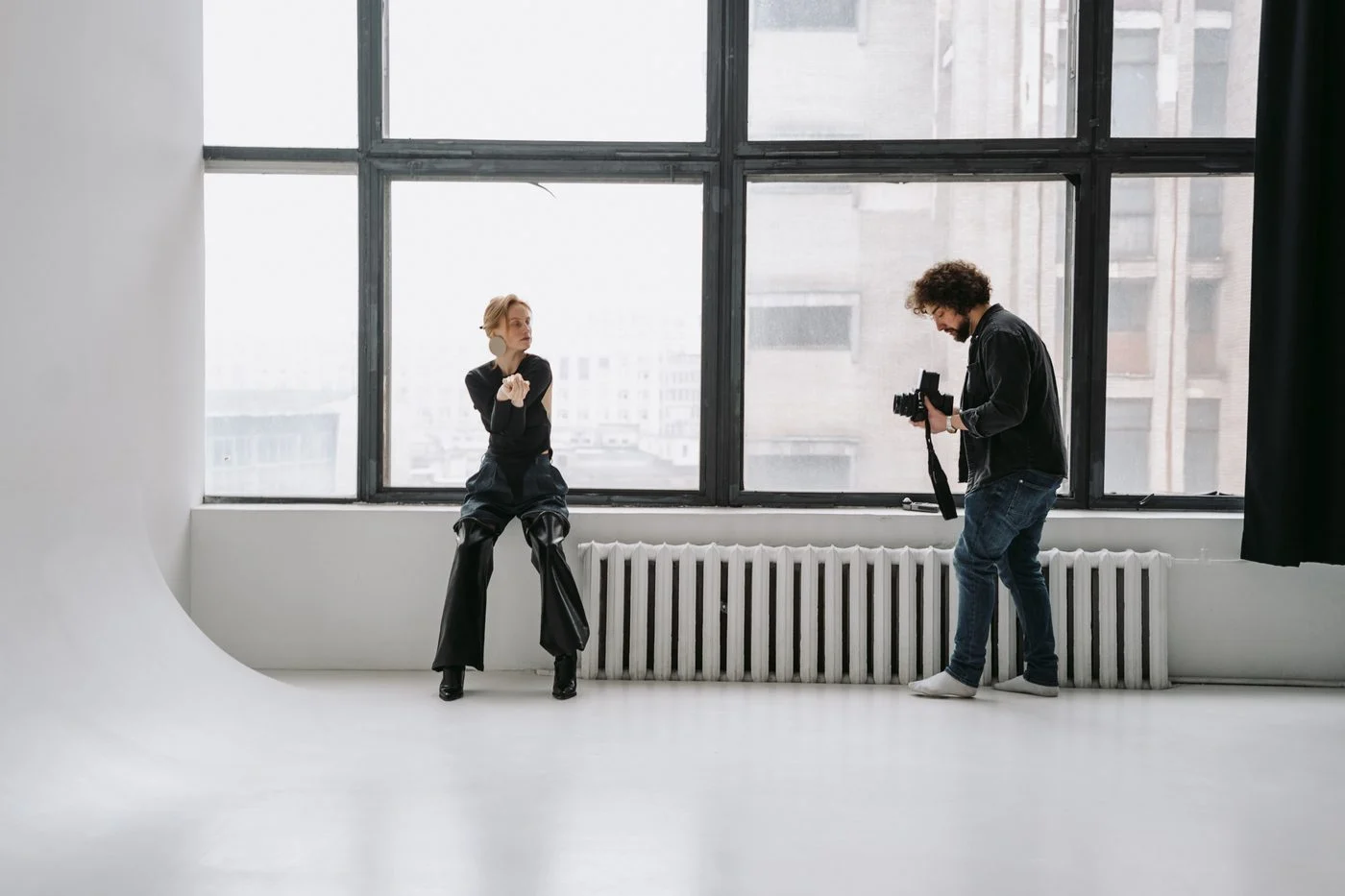 A model seated by large studio windows while a photographer adjusts his camera during a fashion shoot